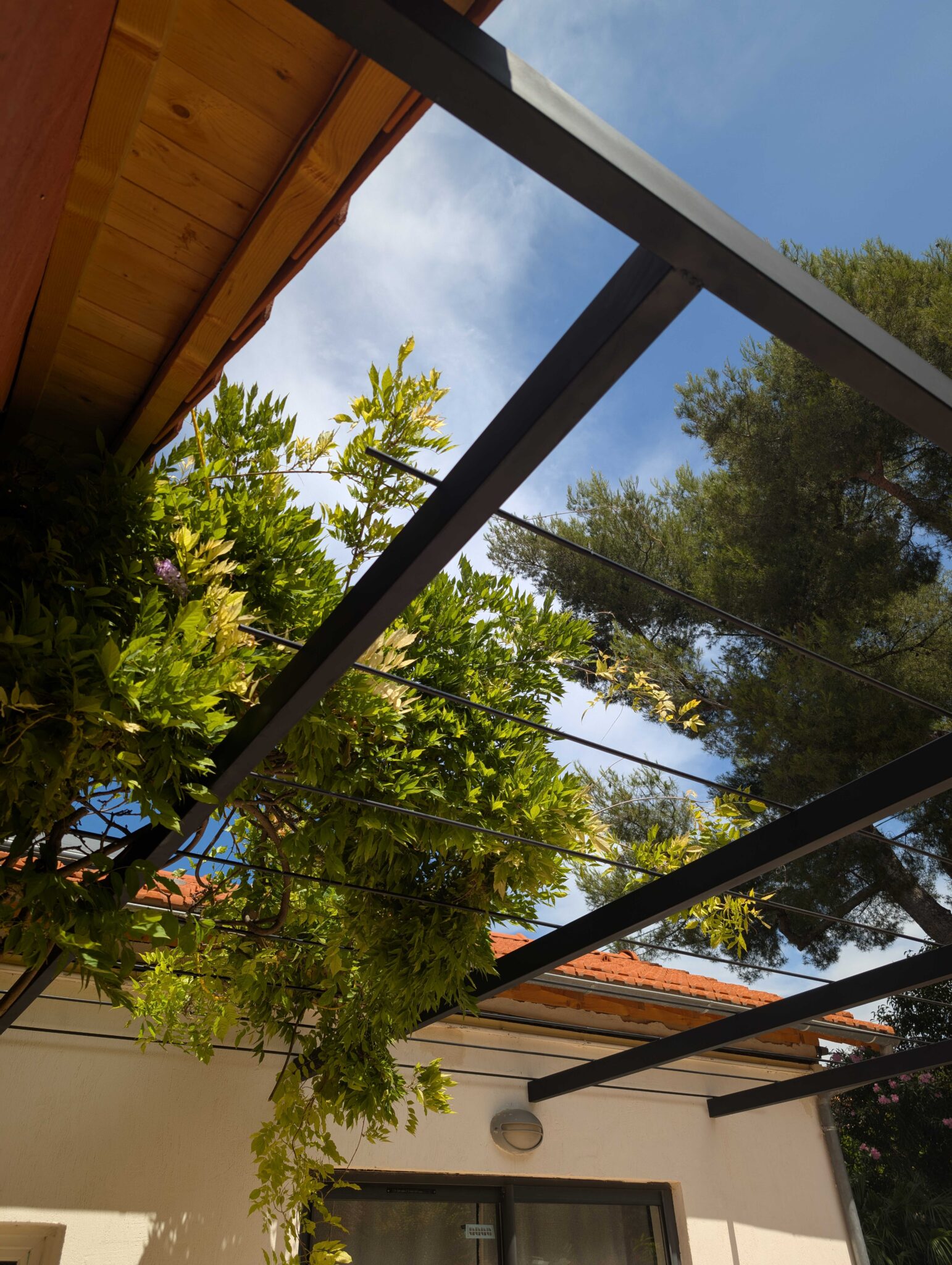 Pergola en acier noir au-dessus d’une terrasse pavée, destinée à recevoir des plantes grimpantes type glycine.