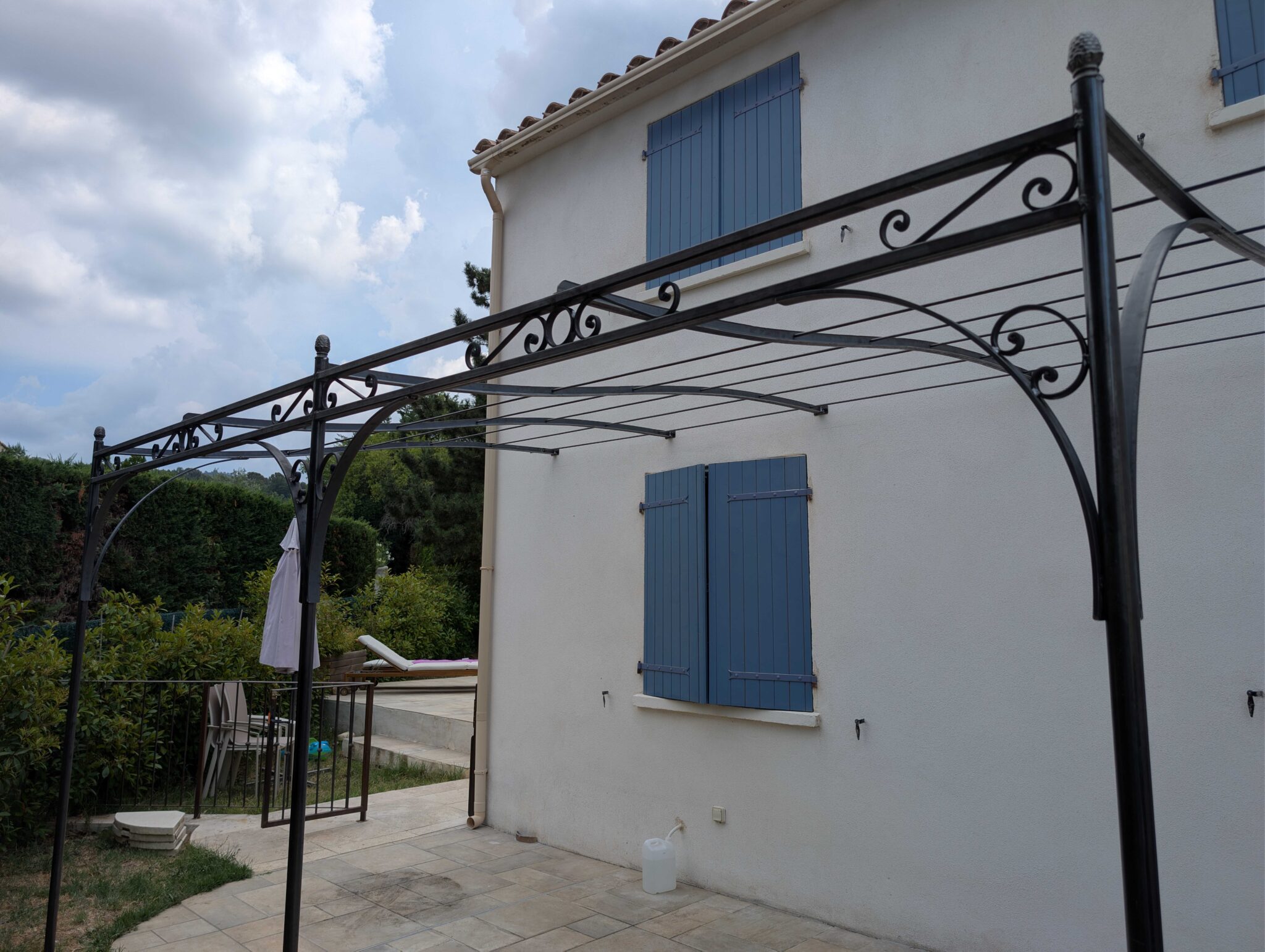 Détail d'une pergola en fer forgé en acier brut avec volutes décoratives et traverses en T.