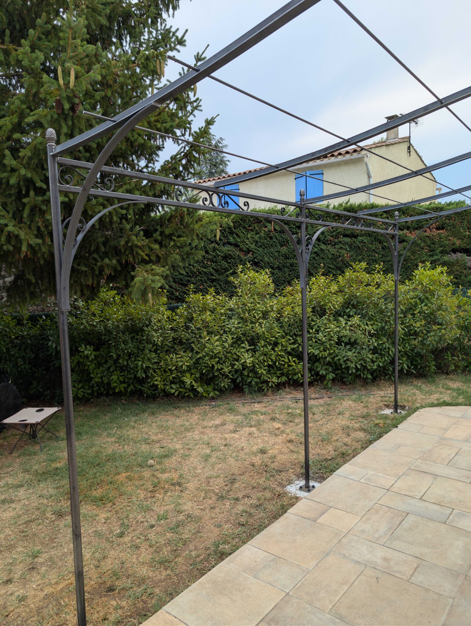 Vue d'ensemble d'une pergola en fer forgé en acier brut avec poteaux ronds et structure décorative.